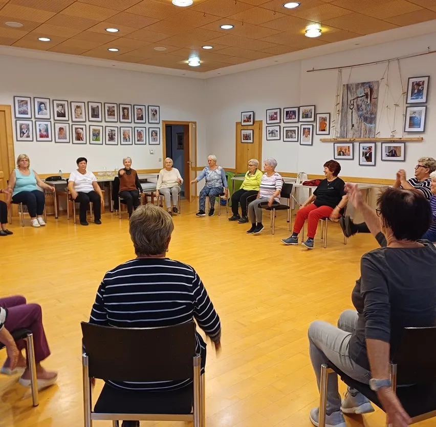 Eine Gruppe älterer Frauen sitzt im Kreis in einem Raum. Sie tragen Freizeitkleidung und scheinen in ein Gespräch vertieft zu sein.
