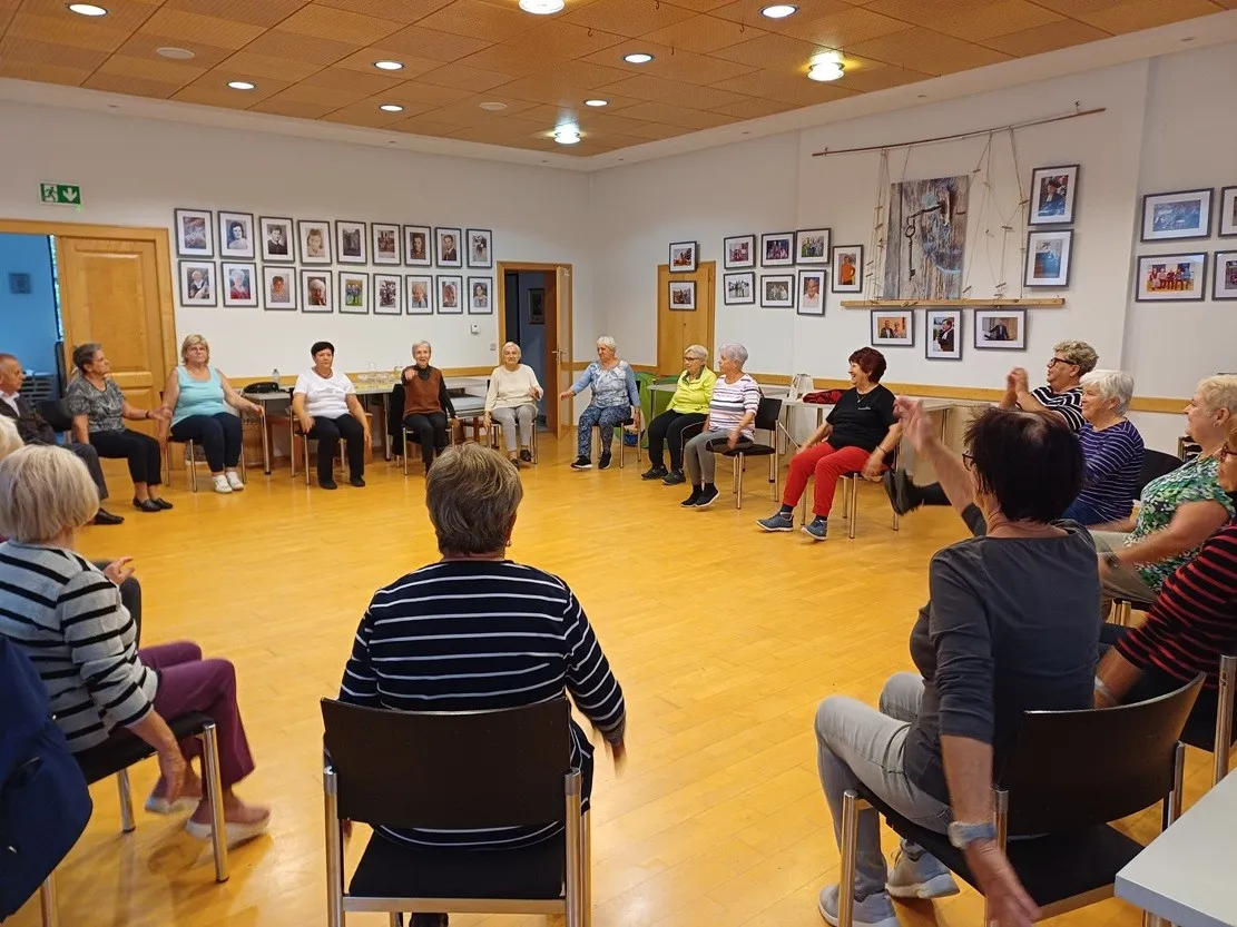Eine Gruppe älterer Frauen sitzt im Kreis in einem Raum. Sie tragen Freizeitkleidung und scheinen in ein Gespräch vertieft zu sein.