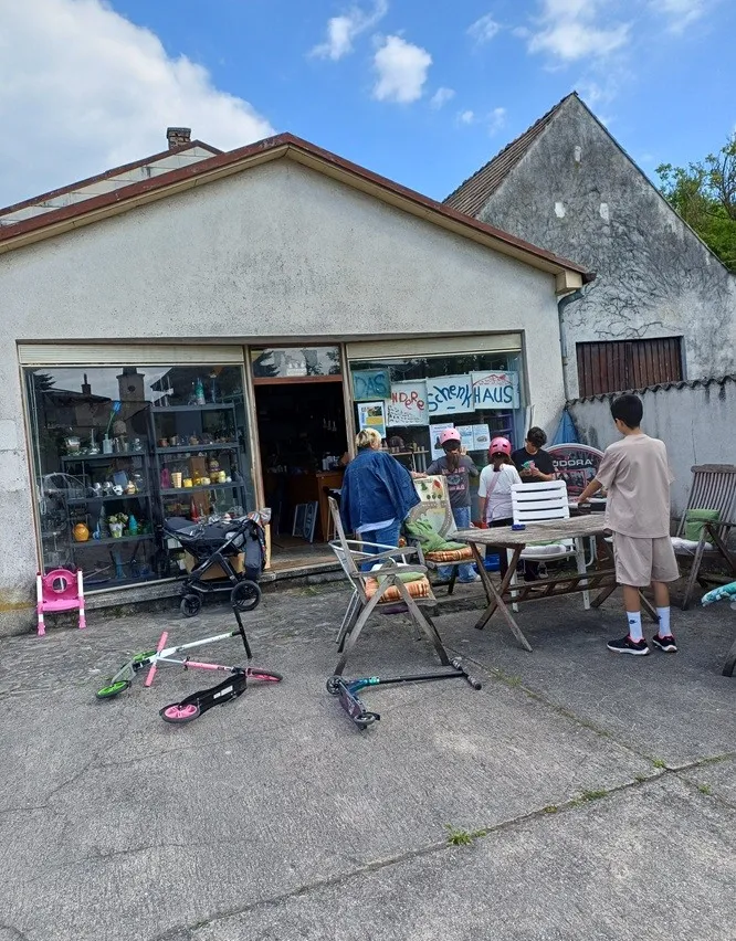 Eine Gruppe von Kindern und Erwachsenen versammelt sich vor einem Geschäft mit einer Glastür. Das Geschäft hat ein Schild, das 'Steakhaus' steht. Ein Kinderwagen, Stühle und ein Tisch sind draußen aufgestellt. Die Kinder tragen Helme.