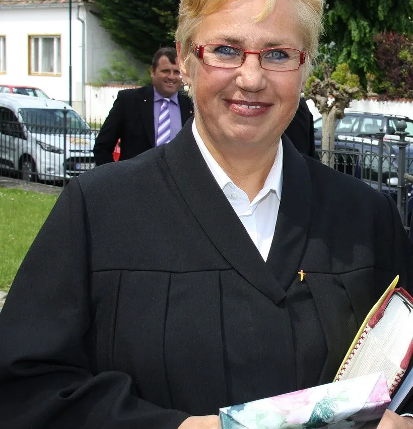 Eine Frau in einem schwarzen Gewand und mit Brille lächelt und hält ein Buch in den Händen. Hinter ihr steht ein Mann in Anzug und Krawatte, und ein Auto ist vor einem Gebäude geparkt.