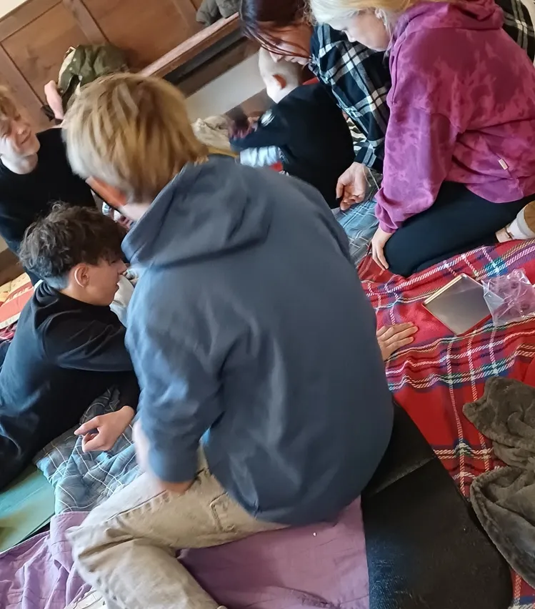 Eine Gruppe von Kindern sitzt auf einer Decke auf dem Boden, wahrscheinlich in einem Klassenzimmer. Sie schauen sich etwas auf dem Boden an.