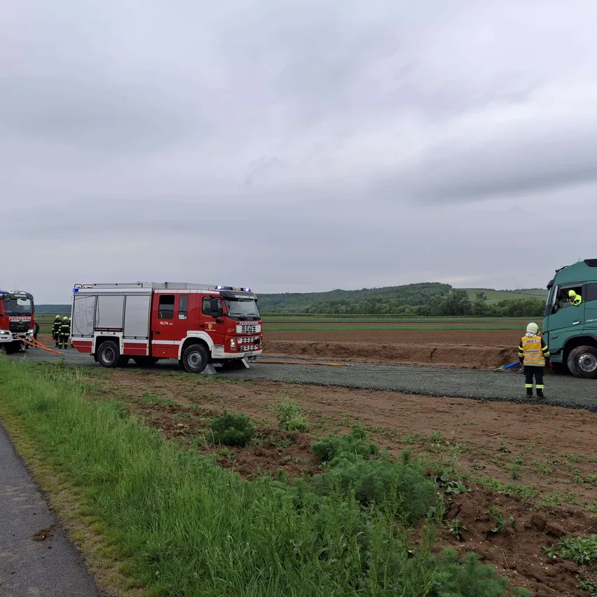 Zwei Feuerwehrfahrzeuge und ein grüner Lastwagen sind am Straßenrand geparkt. Eine Person steht neben dem grünen Lastwagen.