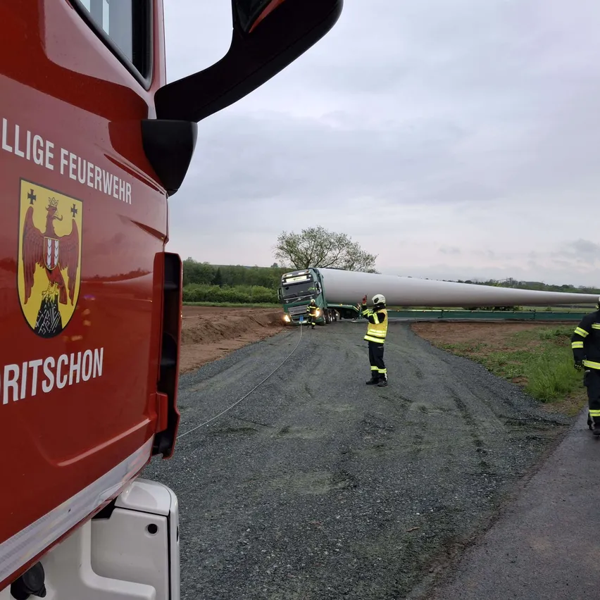Ein Feuerwehrwagen mit der Aufschrift 'Ritschon' auf der Seite steht auf einer Schotterstraße. In der Nähe steht eine Person in einer reflektierenden Sicherheitsweste neben einem LKW, der ein langes, zylindrisches Objekt transportiert, möglicherweise eine Windturbinen-Blatt.
