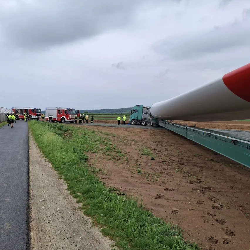 Ein großes weißes und rotes Windturbinenblatt wird auf einem Lastwagen auf einem unbefestigten Weg transportiert. Feuerwehrleute und Arbeiter stehen in der Nähe.