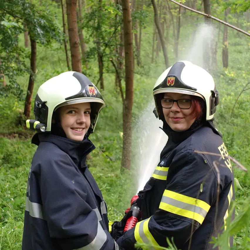 Zwei Feuerwehrleute in Uniform stehen in einem Wald, einer hält einen Feuerlöscher. Sie tragen beide Helme und Brillen. Hinter ihnen sprudelt ein Baumstamm Wasser.