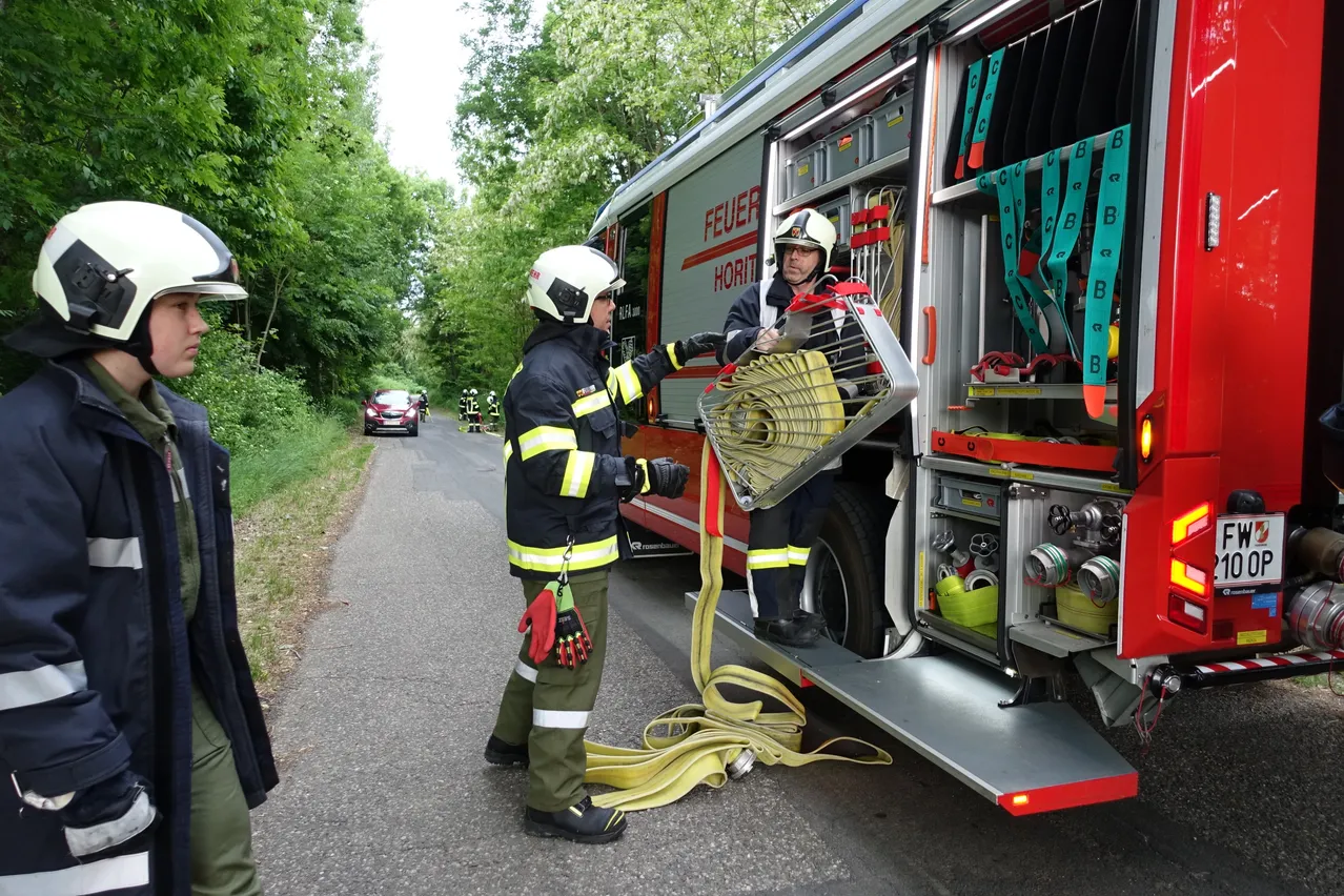 Zwei Feuerwehrleute in voller Montur verlassen einen Feuerwehrwagen am Straßenrand. Einer hält einen Feuerlöscher, während der andere eine Feuerwehrschläuche auszieht.