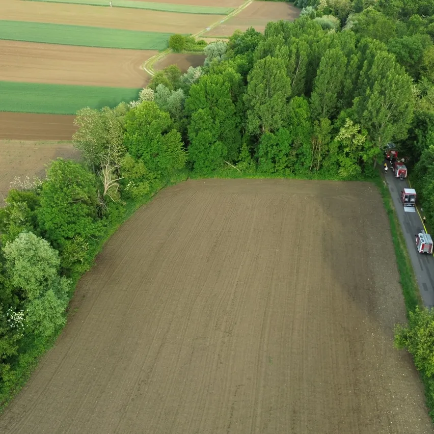 Eine Luftaufnahme eines großen Ackerlandes, umgeben von grünen Bäumen und Büschen. Fahrzeuge fahren auf der Straße rechts.