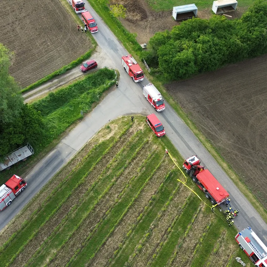 Eine Luftaufnahme mehrerer Feuerwehrfahrzeuge auf einer Straße, wobei eines eine Schlange zieht. Es stehen Menschen an der Seite, und ein rotes Auto ist in der Nähe. Die Straße ist von Feldern und grünem Gras umgeben.