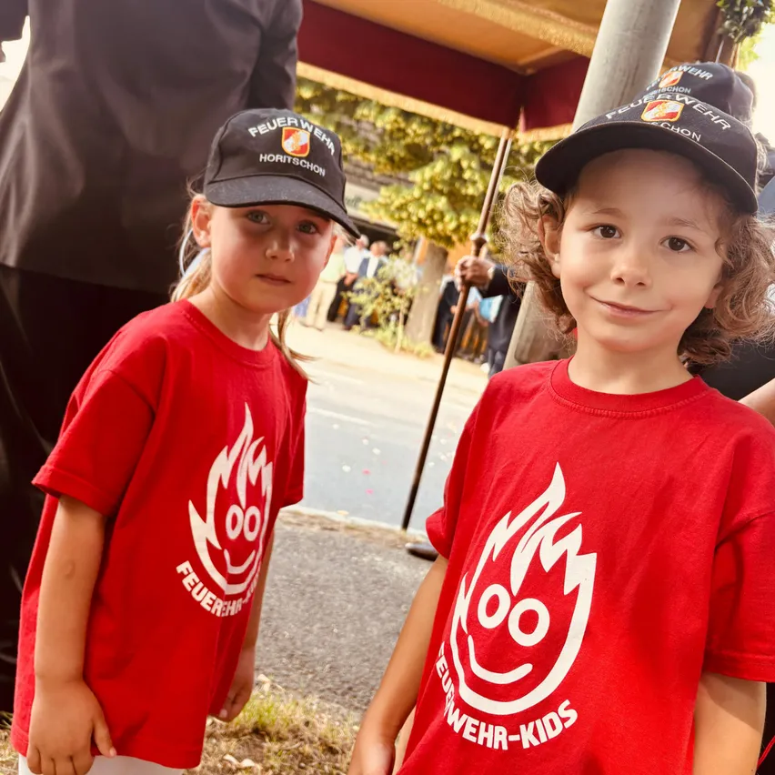 Zwei Kinder in roten T-Shirts mit weißen Logos und Mützen mit Text. Sie stehen draußen, mit einer Person, die einen Stock in der Hand hält, hinter ihnen.