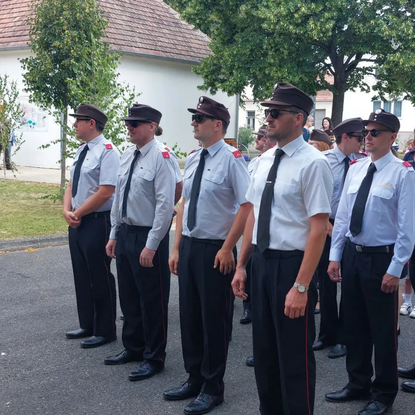 Eine Gruppe uniformierter Männer steht in einer Reihe, möglicherweise bei einer Parade. Sie tragen Hüte, Krawatten und haben Abzeichen auf der Brust. Hinter ihnen stehen Bäume und Gebäude.