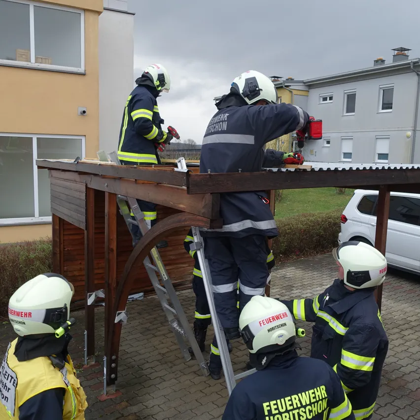 Eine Gruppe Feuerwehrleute arbeitet mit Leitern und Werkzeugen an einer Holzstruktur. Sie tragen Helme und Uniformen mit gelben und schwarzen Streifen.