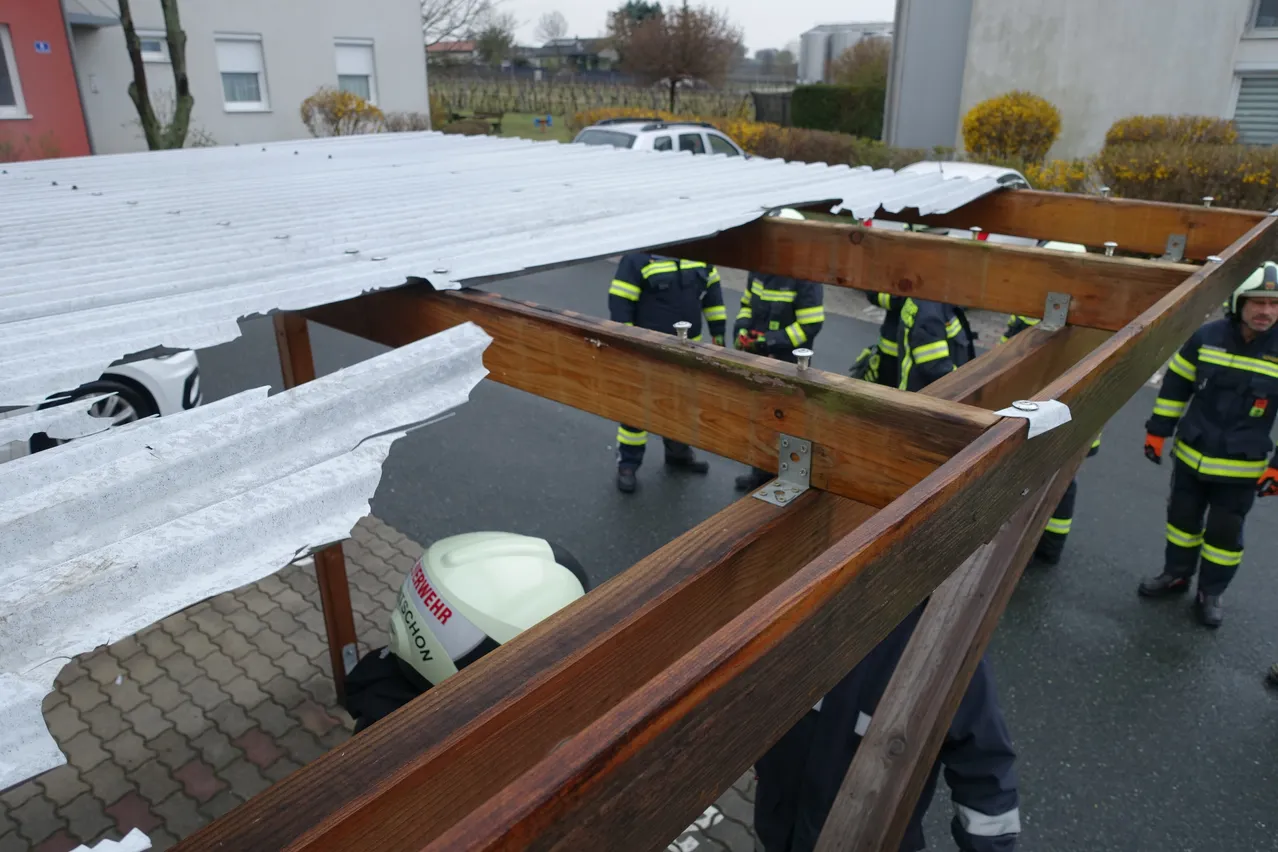 Feuerwehrleute arbeiten an einem beschädigten Dach, wobei einige Teile entfernt wurden. Im Vordergrund ist ein weißer Helm sichtbar.