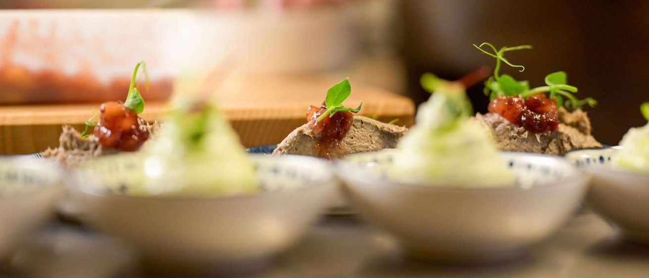 Ein Koch bereitet Gerichte mit roten und grünen Garnierungen auf einem Tresen vor. Im Vordergrund befinden sich drei Teller mit kunstvoll angerichteten Speisen. Im Hintergrund sind die Hände des Kochs verschwommen, was Bewegung anzeigt.