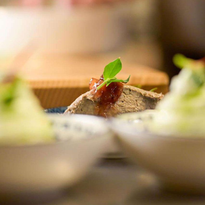 Ein Koch bereitet Gerichte mit roten und grünen Garnierungen auf einem Tresen vor. Im Vordergrund befinden sich drei Teller mit kunstvoll angerichteten Speisen. Im Hintergrund sind die Hände des Kochs verschwommen, was Bewegung anzeigt.
