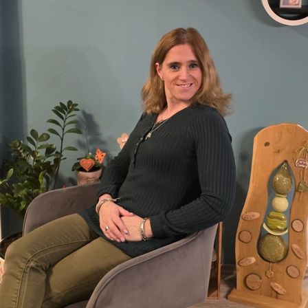 Eine Frau sitzt auf einem Stuhl und lächelt in die Kamera. Hinter ihr sind drei kreisförmige Regale an der Wand befestigt, die Figuren und ein Fotorahmen zeigen. Pflanzen befinden sich auf beiden Seiten von ihr.