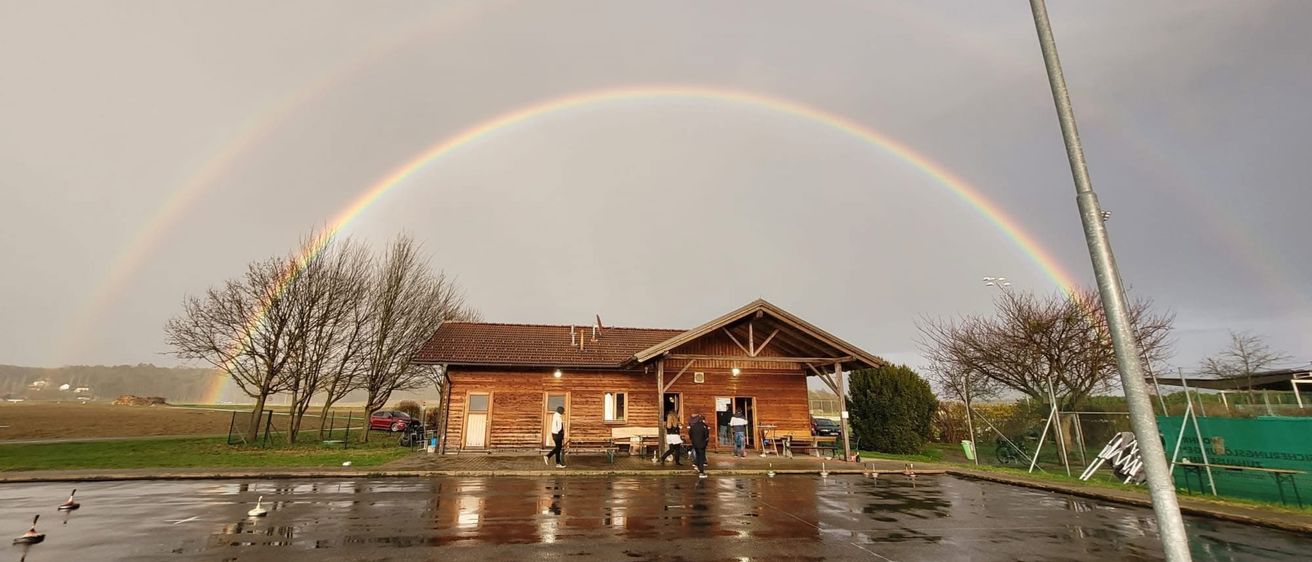 Ein Regenbogen spannt sich über ein kleines Gebäude, vor dem Menschen stehen. Die Szene ist nass, mit einem geparkten Auto in der Nähe und Bäumen an den Seiten.