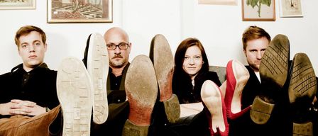 Fünf Personen sitzen auf einer Couch mit hochgelegten Füßen auf einem Tisch, in Freizeitkleidung. Sie sind umgeben von gerahmten Bildern an einer weißen Wand.