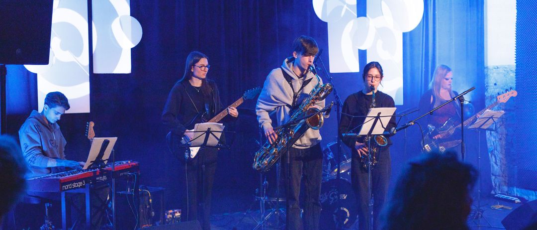 Vier Personen auf der Bühne mit Musikinstrumenten. Zwei von ihnen spielen Saxophon, einer spielt Gitarre und einer spielt Klavier. Sie treten vor einem blauen Hintergrund mit einem weißen Logo auf.