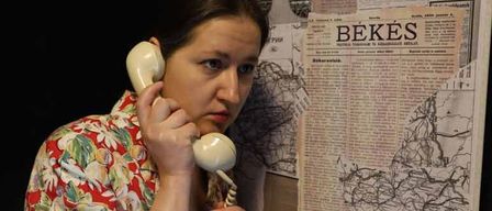 A woman in a floral dress stands behind a phone booth, holding the receiver with a serious expression. Behind her, a map and newspapers are pinned to the wall.