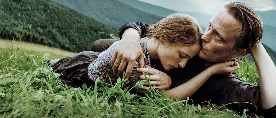 Ein Mann und eine Frau liegen sich umarmend auf einem Grasfeld. Der Mann hat seinen Kopf auf der Brust der Frau. Sie sind wahrscheinlich in einem Wald. Dahinter ist ein Berg.