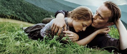 Ein Mann und eine Frau liegen sich umarmend auf einem Grasfeld. Der Mann hat seinen Kopf auf der Brust der Frau. Sie sind wahrscheinlich in einem Wald. Dahinter ist ein Berg.