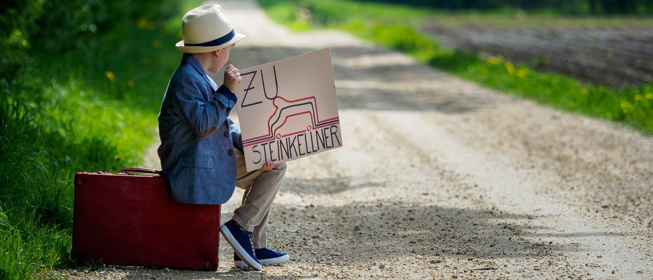 Ein junger Junge in einem Hut sitzt auf einem roten Koffer auf einem Schotterweg und hält ein Pappschild mit der Aufschrift 'ZU STEINKELLNER' in der Hand.