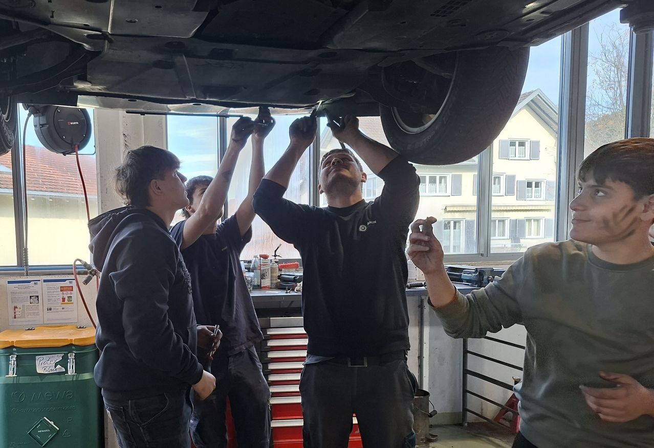 Vier Männer arbeiten in einer Werkstatt an einem Auto, einer hält ein Werkzeug und arbeitet unter dem Auto, während die anderen zusehen.