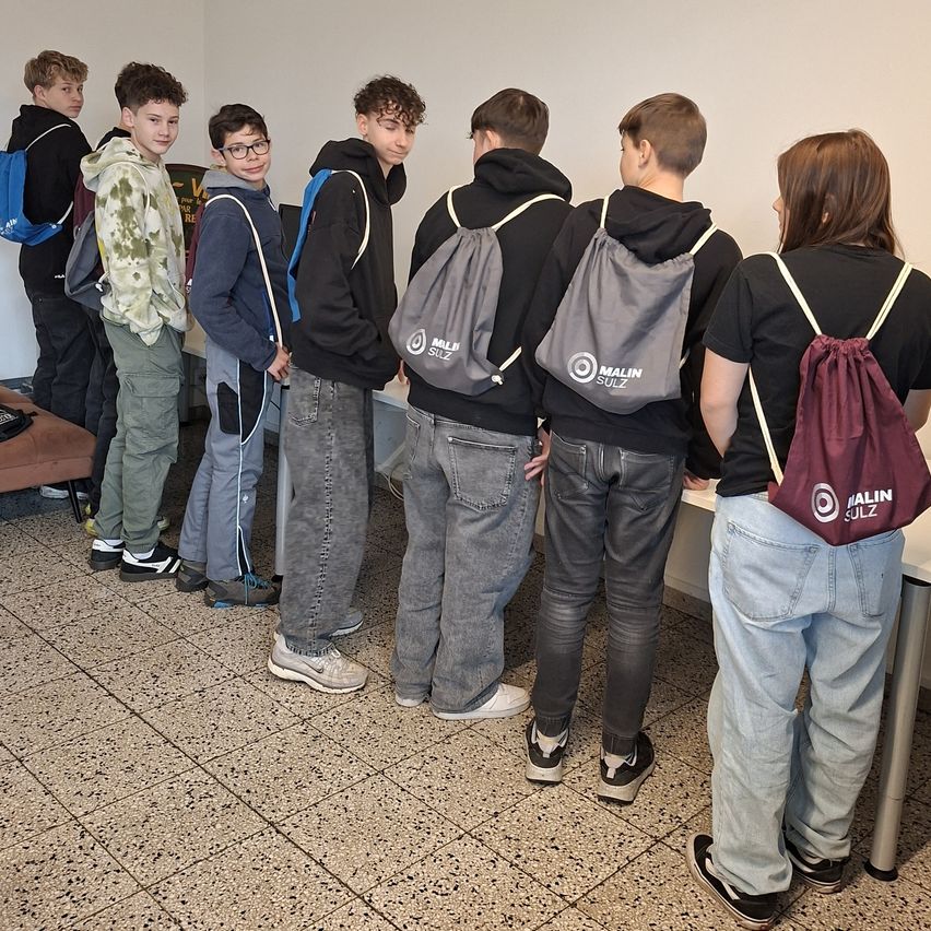 Eine Gruppe junger Personen, lässig gekleidet, steht in einer Reihe, mit dem Rücken zur Kamera, an einer weißen Wand. Einige tragen Rucksäcke.