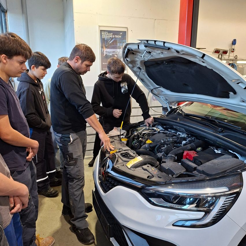 Eine Gruppe von Menschen beobachtet einen Mann, der in einer Garage an einem Motor eines weißen Autos arbeitet.