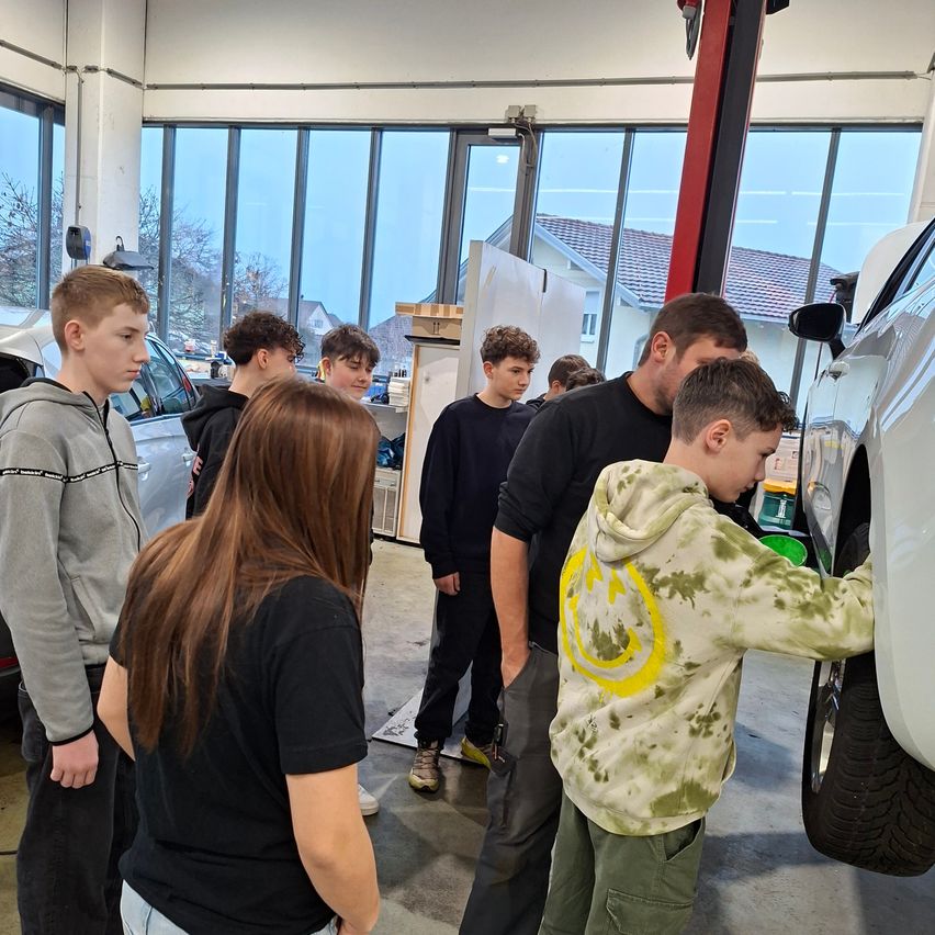 In einer Autowerkstatt beobachtet eine Gruppe von Menschen, wie ein Mann das Rad eines Autos repariert. Zwei Autos sind geparkt, und ein Auto ist angehoben. Die Werkstatt hat große Fenster und ein Haus im Freien.