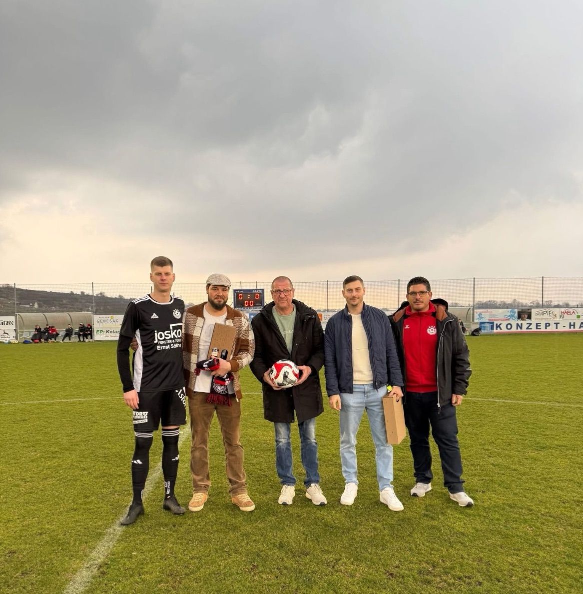 Fünf Männer stehen auf einem Fußballfeld und posieren mit einem Fußball und Pokalen. Sie sind für kaltes Wetter gekleidet. Hinter ihnen trennt ein Zaun das Feld vom wolkigen Himmel.