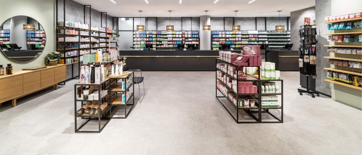 Eine gut beleuchtete Apotheke mit modernen Regalen und ausgestellten Produkten. Helle Deckenleuchten und ein sauberes Layout.