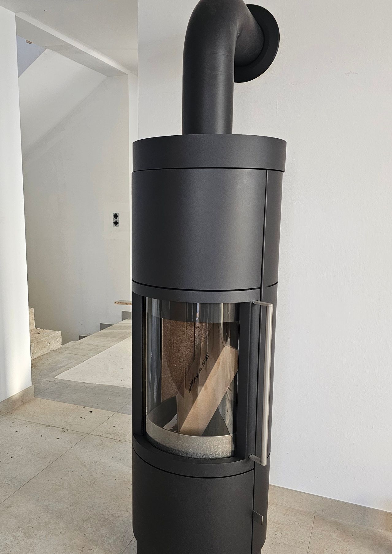 Ein schwarzer Holzofen mit Glastür steht an einer weißen Wand neben einem weißen Boden und einer Treppe.