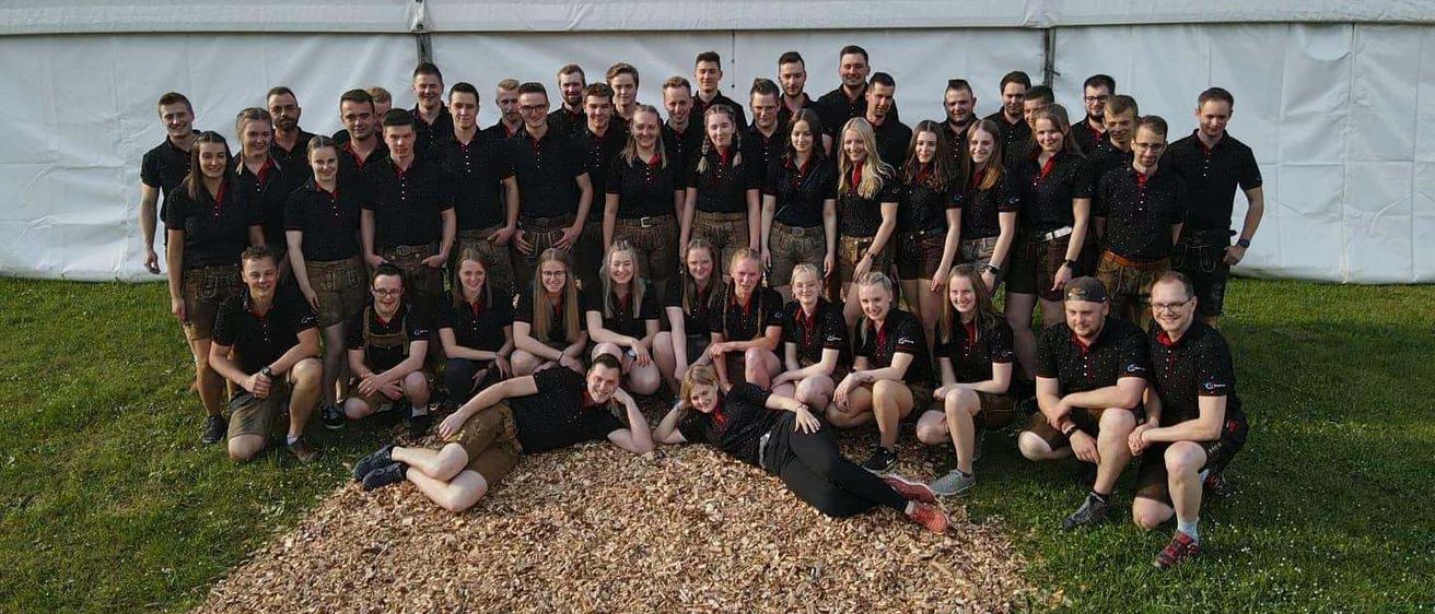 Eine Gruppe junger Leute in Kilts und schwarzen Shirts posiert für ein Foto vor einem Zelt mit einem gelben und blauen Schild, das Lungauer Zeltverleih steht.