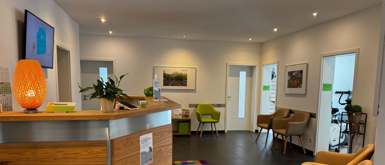 Ein moderner Büroempfangsbereich mit einer hölzernen Theke, einem Topfpflanzen, einem bunten Teppich und mehreren Stühlen. Es gibt gerahmte Bilder an der Wand und einen Monitor auf der linken Seite.