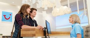 Drei Menschen sind in einem Gebäude mit weißen Wänden und Decken. Zwei Menschen lächeln und unterhalten sich mit der dritten Person. Sie stehen vor einem Tresen mit einem Computer.