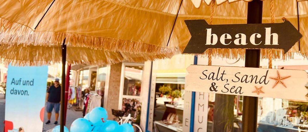 Ein Strandgeschäft mit einem Schild 'Willkommen am Strand' und einem weiteren, das 'Salz, Sand & Meer' sagt. Es gibt einen Tisch mit blauen Luftballons, einem Mann in einem schwarzen T-Shirt und einem geparkten Fahrrad in der Nähe.