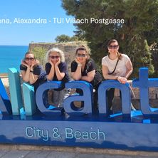 Vier Frauen posieren vor einem Schild von Alicante City and Beach mit Küstenhintergrund.