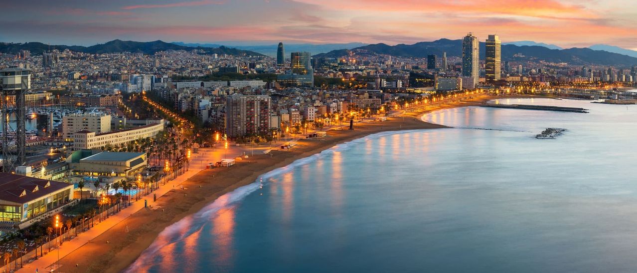 Der Skyline einer Küstenstadt bei Sonnenuntergang mit Hochhäusern und einem breiten Strand mit beleuchteten Straßenlaternen.