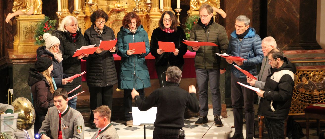 Ein Chor von Erwachsenen steht vor einem Altar in einer Kirche, hält rote Liederbücher in der Hand, mit einem Dirigenten und Musikinstrumenten. Der Chor tritt in einer Kirche auf.