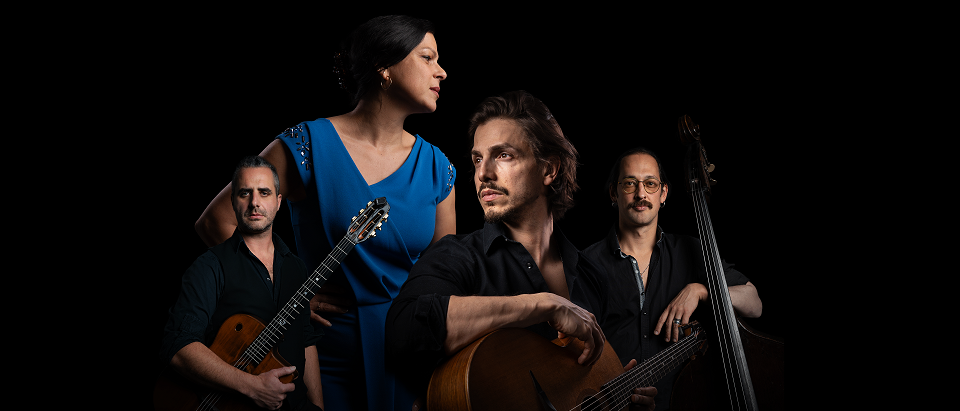 Vier Musiker in einer Gruppenaufnahme. Drei Männer und eine Frau, alle in Schwarz gekleidet. Die Frau trägt ein blaues Kleid. Sie halten Gitarren und einen Bass.