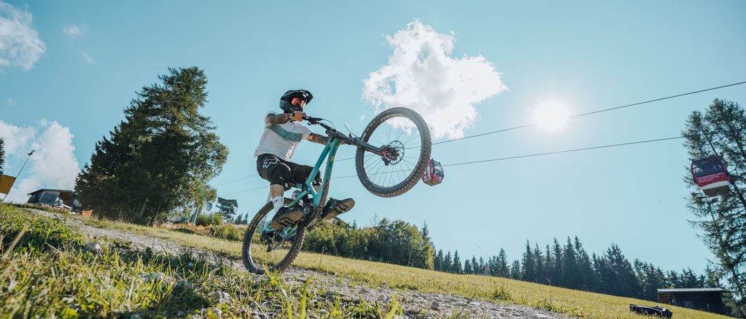 Ein Mountainbiker fährt auf einem Trail mit einem Rad in der Luft. In der Ferne sind Bäume und eine Seilbahn zu sehen.