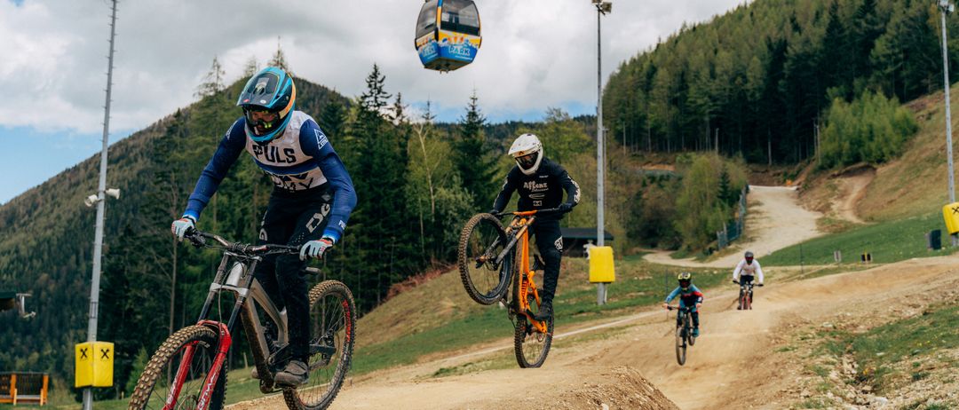 Eine Gruppe von Radfahrern fährt auf einer Dirt-Piste, mit einer Seilbahn darüber. Einer springt auf dem Fahrrad.