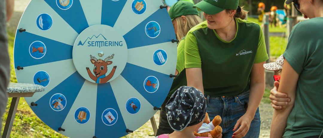 Eine Frau mit grüner Mütze und grünem Shirt steht neben einem großen Drehrad mit der Aufschrift Semmering, mit einem Kind, das ein Kuscheltier hält. Hinter ihnen sind andere Leute und ein Spielplatz.