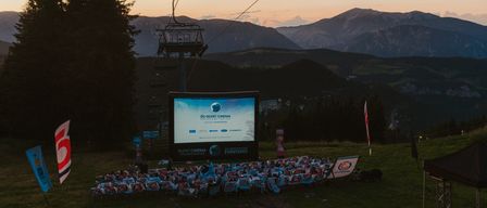 Stummfilm-Veranstaltung am Berghang mit einer großen Leinwand, umgeben von Leuten auf Stühlen. Oben ist eine Skiliftstation.