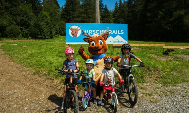 Vier Kinder, ein Maskottchen und Fahrräder, alle versammelt vor dem Hirschtrails-Schild in einer malerischen Gegend.
