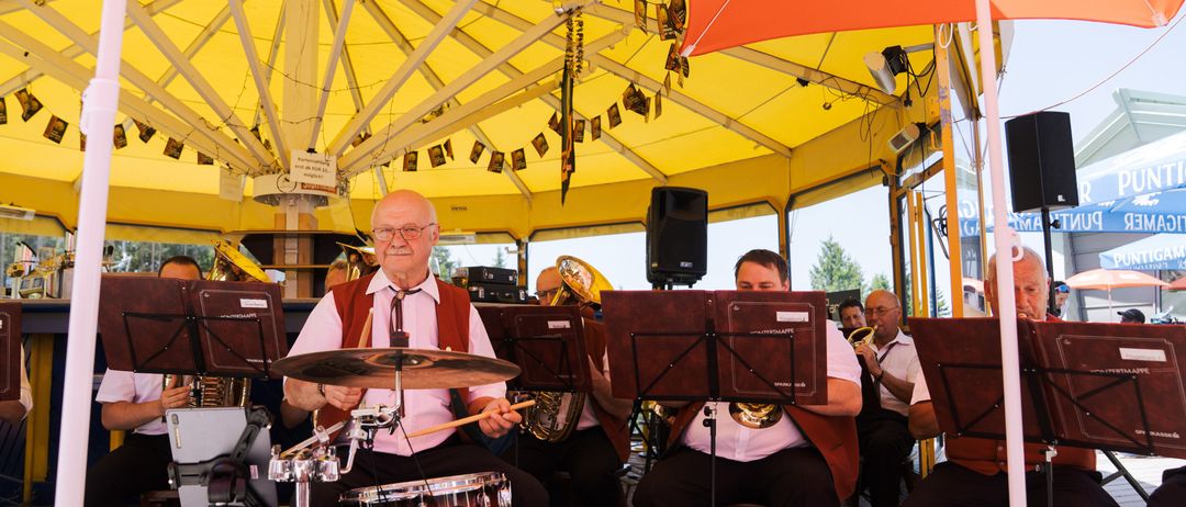 Ein älterer Mann spielt die Trommeln unter einem großen gelben Zelt, während eine Brassband im Hintergrund spielt.