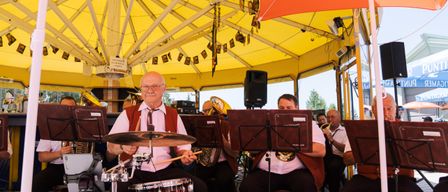An older man plays the drums under a large yellow tent with a brass band performing in the background.