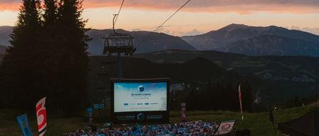 Eine große Outdoor-Filmleinwand ist in einem Berggebiet mit einem Skilift im Hintergrund aufgebaut. Die Leinwand zeigt einen Filmtitel, und viele Menschen sitzen vor ihr in Stühlen.