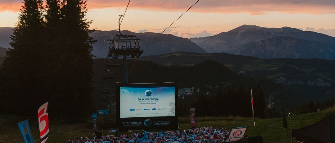 Eine große Outdoor-Filmleinwand ist in einem Berggebiet mit einem Skilift im Hintergrund aufgebaut. Die Leinwand zeigt einen Filmtitel, und viele Menschen sitzen vor ihr in Stühlen.